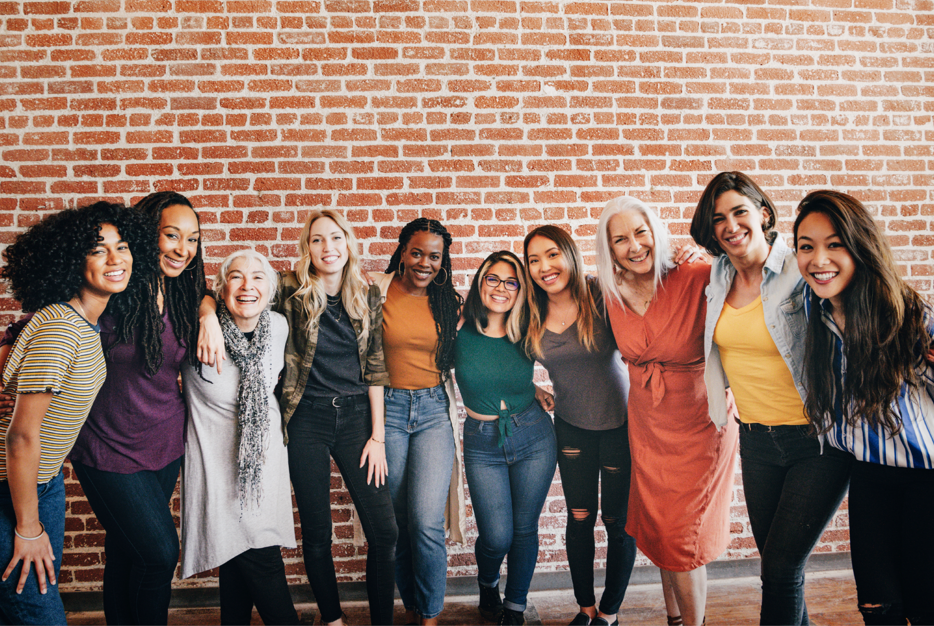 a row of smiling women standing in front a brick wall, with arms around each other