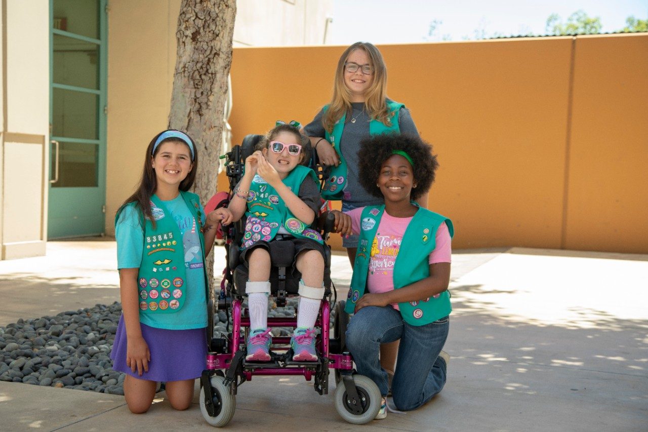 four girl scouts smiling, one is standing, two are kneeling and one is in a wheelchair
